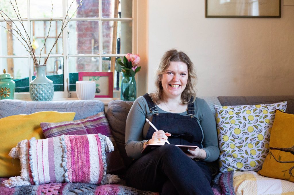 Alex sitting on her sofa with pencil in her hand smiling at the camera 