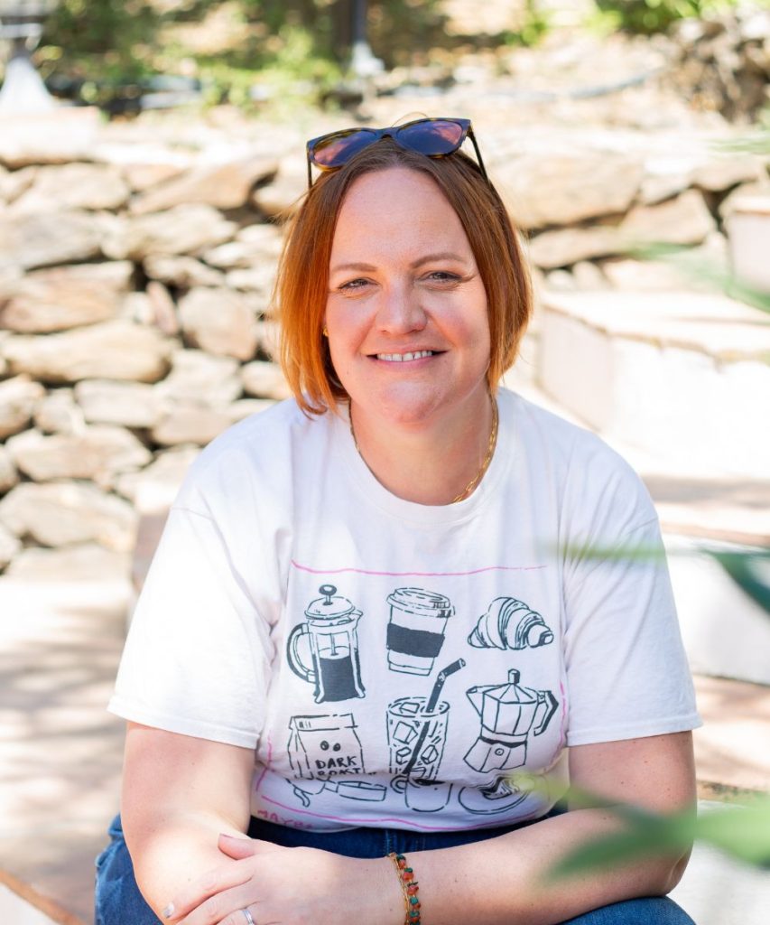 Corinne sitting outside in the sunshine smiling at the camera with sunglasses on her head